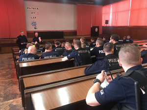 Aula w komendzie miejskiej Policji w Gliwicach. Liczni policjanci w ławkach spisują w notatnikach polecenia prowadzących odprawę.