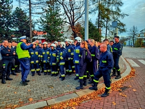 Policjant drogówki szkoli grupę kilkudziesięciu strażaków. Stoją na zewnątrz budynku.