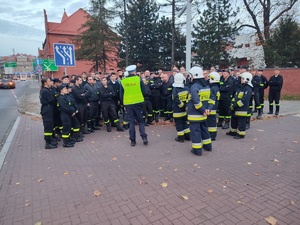 Policjant prowadzi wykład na ulicy dla kilkudziesięciu strażaków. Stoją w pobliżu skrzyżowania.