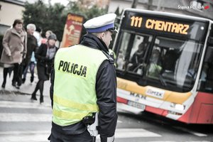 Policjant ruchu drogowego a w tle przejście dla pieszych, autobus komunikacji miejskiej i ludzie odwiedzający cmentarz