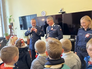 Na pierwszym planie dzieci klasy 1 B ze szkoły podstawowej numer 6 w Gliwicach. w tle dwaj oficerowie policji - maja założone na mundury kotyliony. po prawej uśmiechająca się policjantka - tez posiada kotylion.