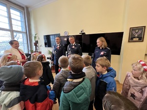 Na pierwszym planie dzieci klasy 1 B ze szkoły podstawowej numer 6 w Gliwicach. w tle dwaj oficerowie policji - maja założone na mundury kotyliony. po prawej uśmiechająca się policjantka - tez posiada kotylion.