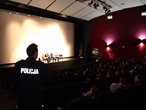 Na zdjęciu widzimy salę kinową. widzimy policjanta patrzącego na widownie - gdzie siedzą młodzi ludzie.