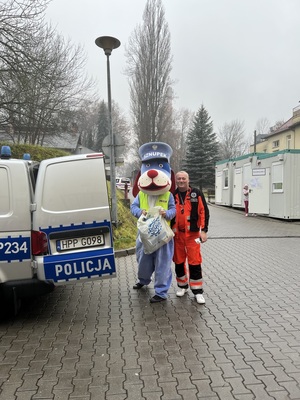 Na zdjciu widzimy policyjny radiowóz a przy nim policyjna maskotka - Pies Sznupek (osoba w kostiumie) obok stoi ratownik medyczny.