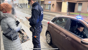 Na zdjęciu umundurowany policjant kontroluje pieszą. Po prawej na ulicy stoi policyjny radiowóz a w nim policjant.