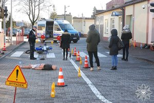 Szkolenie - inscenizacja miejsca wypadku rowerzysty. Na asfalcie lezy manekin, rower. policjant tłumaczy, kilka osób słucha.