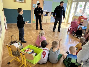 Na zdjęciu policjant i policjantka rozmawiają z dziećmi przedszkolnymi. Policjanci stoją przed siedzącymi na podłodze maluchami.