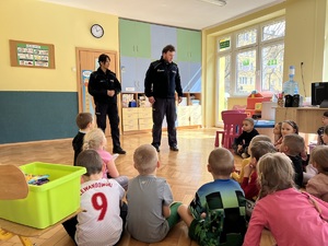 Na zdjęciu policjant i policjantka rozmawiają z dziećmi przedszkolnymi. Policjanci stoją przed siedzącymi na podłodze maluchami.