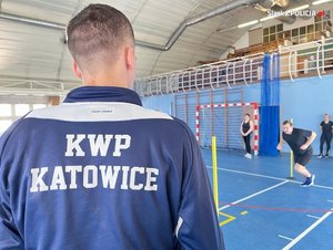 Na zdjęciu widzimy instruktora sportowego w koszulce z napisem KWP Katowice stoi plecami do kadru, w tle mężczyzna wykonujący test sportowy.