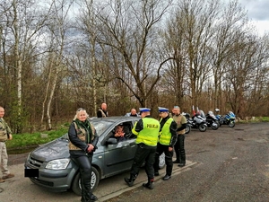 Na zdjęciu widzimy kontrolę samochodu osobowego przez dwóch policjantów. Towarzysza im cywile w strojach motocyklowych. W tle zaparkowane motocykle.