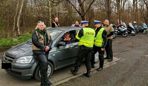 Na zdjęciu widzimy kontrolę samochodu osobowego przez dwóch policjantów, towarzysza im osoby cywilne - motocykliści. w tle zaparkowane różne motocykle.