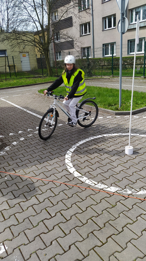 Na zdjęciu widzimy dziewczynkę na rowerze, która przejeżdża przez skrzyżowanie o ruchu okrężnym - tak zwane rondo.