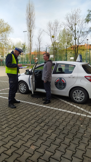 Na zdjęciu widać, jak policjant wraz uczestnikiem konkursu przygotowują się do losowania pytań egzaminacyjnych. Stoją obok pojazdu.