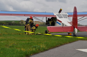 Na tle płyty lotniska na skraju pasa startowego jest widoczny niewielki, osobowy samolot w kolorach czerwono-biało-niebieskim, stojący częściowo na trawie. W centralnym punkcie zdjęcia są widoczni strażacy rozmawiający z ofiarą wypadku lotniczego leżącą pod samolotem, dodatkowo widać jak jeden ze strażaków próbuje wyciągnąć kolejną osobę z samolotu. W dolnej części zdjęcia widać rozciągnięty żółty wąż strażacki.