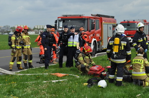 Na pierwszym planie na tle płyty aeroklubu gliwickiego oraz dwóch wozów bojowych straży pożarnej są widoczne wszystkie służby ratownicze – strażacy, ratownicy medyczni oraz policja, łącznie około 12 osób, każdy wykonuje zadania przydzielone przez widocznego w centralnym punkcie dowódcę działań – Naczelnika Wydziału Sztab Policji w Gliwicach. W prawym dolnym rogu widać dodatkowo częściowo leżącą na noszach ratunkowych jedną z ofiar wypadku.