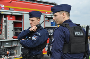 Na tle wozu bojowego straży pożarnej widzimy dwóch funkcjonariuszy Policji z Wydziału Sztab Komendy Miejskiej Policji w Gliwicach – ubrani są w umundurowanie ćwiczebne tak zwane moro, koloru granatowego, na głowie mają furażerki, oraz mają na sobie kamizelki taktyczne. Jeden z Policjantów prowadzi rozmowę za pośrednictwem stacji nasobnej.