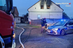 Policyjny radiowóz oraz wóz strażacki na tle budynku w którym ugaszono pożar. Widzimy policjanta i strażaka oraz osobę cywilną.