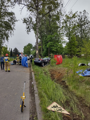 Zdjęcie z oddali pokazujące miejsce zdarzenia, widać drzewom rozbity samochód i wielu funkcjonariuszy krzątających się wokół.