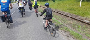 Zdjęcie przedstawia jadących rowerzystów (zdjęcie zrobione od tyłu) po drodze publicznej przy torach tramwajowych. Wśród kolarzy można zauważyć dzieci z rodzicami, młodzież i osoby dorosłe.