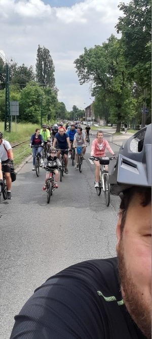 Zdjęcie przedstawia jadących rowerzystów (zdjęcie zrobione od przodu) po drodze publicznej przy torach tramwajowych. Wśród kolarzy można zauważyć dzieci z rodzicami, młodzież i osoby dorosłe.