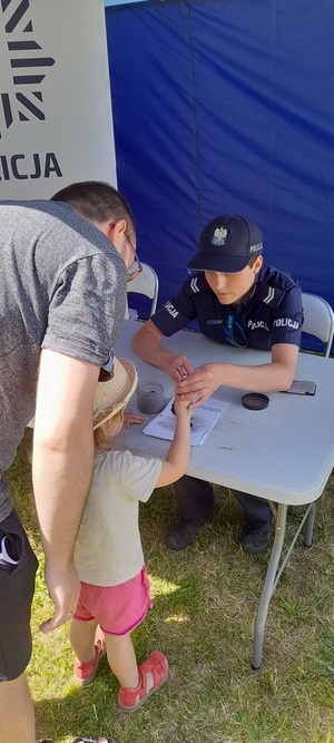 Zdjęcie przedstawia uczestnictwo Policji na VII Gliwickim Święcie Rodziny organizowanego w Gliwicach na terenie Parku Chopina. Na zdjęciu jest widoczne wnętrze namiotu policyjnego, gdzie funkcjonariuszka gliwickiej policji odbijająca dłonie na kartce (technika kryminalistyczna) małej dziewczynki z ojcem.