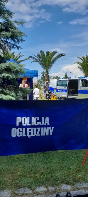 Zdjęcie przedstawia uczestnictwo Policji na VII Gliwickim Święcie Rodziny organizowanego w Gliwicach na terenie Parku Chopina. Na zdjęciu widzimy na pierwszym planie transparent POLICJA OGLĘDZINY, a w tle dwie palmy, oraz po lewej stronie namiot policyjny, a po prawej stronie  pusty  ustawiony bokiem radiowóz oznakowany typu bus.