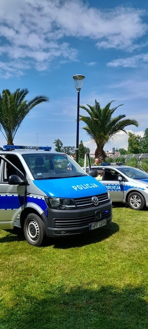 Zdjęcie przedstawia uczestnictwo Policji na VII Gliwickim Święcie Rodziny organizowanego w Gliwicach na terenie Parku Chopina. Na zdjęciu widzimy na pierwszym planie widzimy dwa radiowozy: pierwszy pusty ustawiony bokiem radiowóz oznakowany typu bus, drugi pusty ustawiony bokiem radiowóz oznakowany typu kombi, dodatkowo w tle dwie palmy.