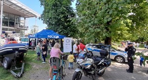 Zdjęcie zrobione podczas pikniku rodzinnego w Gliwicach w Parku Chopina, w tle jest widoczna Palmiarnia. na pierwszym planie widzimy profilaktyczne stanowisku gliwickiej Policji - granatowy namiot z białym stolikiem, koło namiotu jest ustawiony motocykl policyjny, Łódka pontonowa na przyczepce, dwa radiowozy - bus i kombi. dookoła stoiska stoją osoby dorosłe i dzieci, na pierwszym planie widać panią trzymającą dwa rowery.