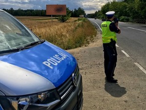 Na zdjęciu widzimy Policjanta w umundurowaniu służbowym, dodatkowo ma założoną kamizelkę odblaskową. Policjant stoi przy radiowozie oznakowanym typu bus przy krawędzi drogi wraz z urządzeniem mierzącym prędkość.