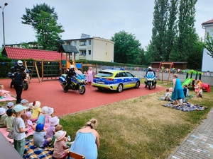 Zdjęcie przedstawia spotkanie dzielnicowych z dziećmi. W centralnym punkcie widzimy radiowóz oznakowany i dwa motocykle oraz umundurowaną policjantkę prowadząca prelekcje.