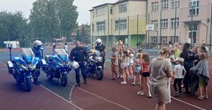 Zdjęcie przedstawia spotkanie dzielnicowych z dziećmi na boisku szkolnym. W centralnym punkcie widzimy trzy motocykle wraz z kierującymi umundurowanymi policjantami oraz umundurowaną policjantkę prowadząca prelekcje.