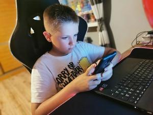 Na zdjęciu widać dziecko w wieku szkolnym, które siedzi na krześle przy biurku i gra na telefonie w grę.