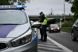 Na zdjęciu widzimy na pierwszym planie przód oznakowanego radiowozu zaparkowanego na poboczu drogi, a dokładnie jego prawa przednią lampę. W oddali widzimy umundurowanego policjanta zapisującego dane w notatniku, oraz mijające go samochody.