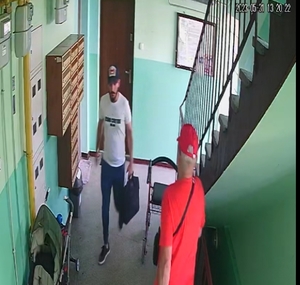 Na zdjęciu widzimy wnętrze klatki schodowej w bloku w Gliwicach, po prawej stronie zamontowane są skrzynki na listy, a po prawej stronie schody prowadzące do góry. Dodatkowo na zdjęciu możemy zobaczyć odwróconego plecami starszego mężczyznę ubranego w czerwoną czapkę z daszkiem oraz czerwoną bluzkę. W centralnej części zdjęcia widzimy osobę poszukiwaną - mężczyznę prawdopodobnie narodowości romskiej z krótką brodą i wąsem. Ubrany jest w niebiesko-białą czapkę z daszkiem, białą bluzkę z krótkim rękawem, długie spodnie typu jeans oraz czarne buty. Mężczyzna trzyma w w lewej ręce torbę na laptopa.