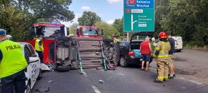 Na zdjęciu widzimy w centralnym miejscu samochód marki Toyota leżący na lewym boku na środku drogi, dookoła pojazdu poruszając się strażacy.