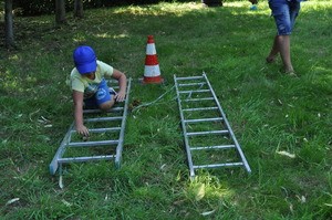 Zdjęcie z pikniku policyjnego, zrobione na terenach zielonych przy Klubie Garnizonowym w Gliwicach, na pierwszym planie widzimy jak dziecko pokonuje kolejną przeszkodę w postaci leżącej drabiny.