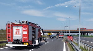 Na zdjęciu widzimy jezdnię autostrady, jest to scena wypadku drogowego. Po lewej znajduje się wóz strażacki, po prawej zamazane miejsce gdzie znajduję się ciało motocyklisty.  w tle policjanci i inne służby.