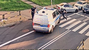 Na zdjęciu widzimy ulicę. Przed sygnalizacja świetlną stoją samochody. Naprzeciw nich stoi bmw, za nim policjny bus z którego wybiegła policjantka i policjant.