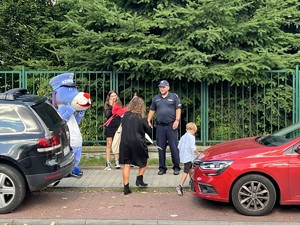 Na zdjęciu widzimy policjanta w towarzystwie policyjnej maskotki - Psa Sznupka rozmawiającego z matka i dzieckiem idącymi do szkoły.