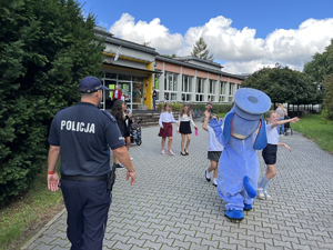 Policjant i policyjna maskotka - Pies Sznupek podchodzą do dzieci stojących przed szkołą.
