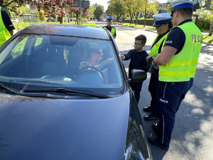 Dwoje policjantów z chłopcem- 8 latkiem wręczają laurkę kierującej siedzącej w samochodzie.
