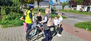Policjantka na chodniku rozmawia z dziećmi ze szkoły podstawowej oraz starsza kobietą prowadząca rower.