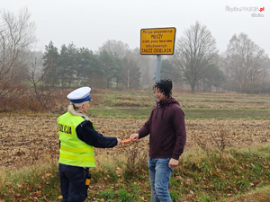 Policjanta wręcza na drodze pieszemu opaskę odblaskową.