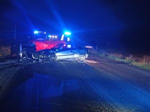 Miejsce wypadku drogowego. Widzimy dwa rozbite samochody osobowe, w tle za parawanem widać pojazdy straży pożarnej i policji.