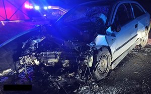 Miejsce wypadku drogowego. Widzimy  rozbite samochody osobowe, w tle za parawanem widać pojazdy straży pożarnej i policji.