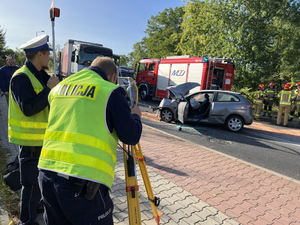 Policjanci mierzą teodolitami miejsce wypadku drogowego. Widać rozbity samochód osobowy.