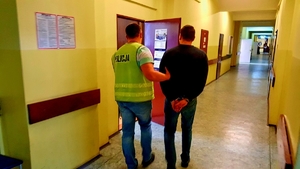 Na zdjęciu korytarz w komendzie miejskiej, policjant wprowadza do pokoju zatrzymanego, który ma skute kajdankami ręce.