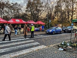 Na zdjęciu widzimy okolice cmentarza, stoją policjanci, spacerują mieszkańcy, widać stragany, po prawej samochody.