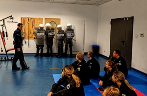 Na zdjęciu widzimy policjanta prezentującego siedzącym na matach uczniom wyposażenie policjantów zabezpieczających imprezy masowe.