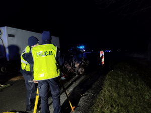 Na zdjęciu dwaj policjanci mierzą urządzeniem typu teodolit miejsce wypadku drogowego.